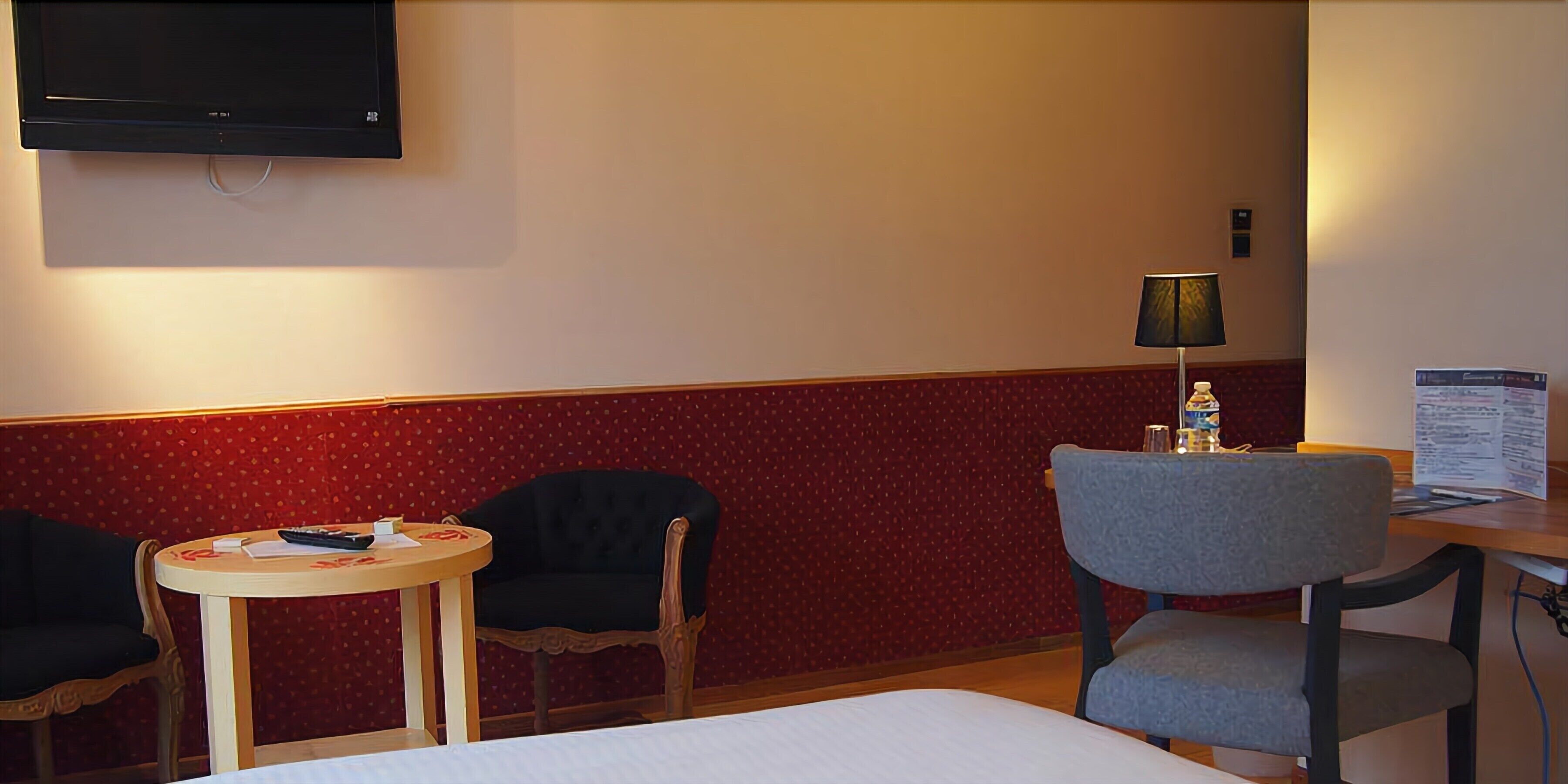 A hotel room with a bed, two chairs, a small table, a lamp, and a TV mounted on the wall.