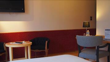 A hotel room with a bed, two chairs, a small table, a lamp, and a TV mounted on the wall.