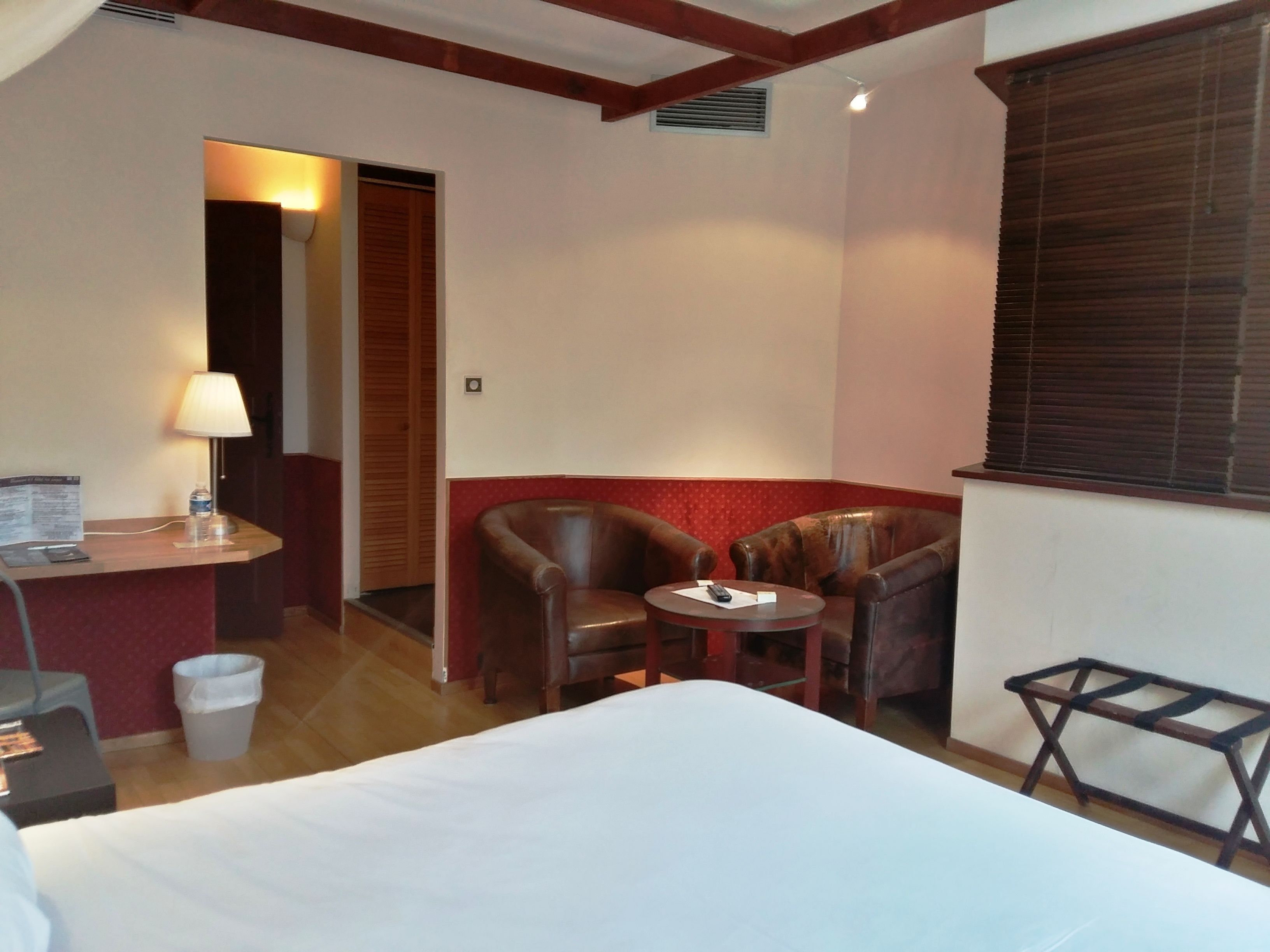 A hotel room with a bed, a desk with a lamp, two leather armchairs, a small table, and a window with blinds.