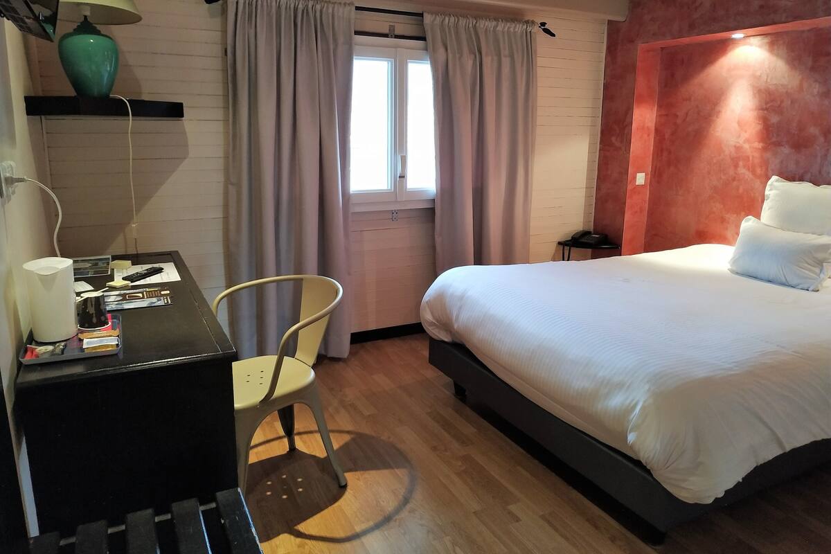 A hotel room with a bed, a desk with a lamp, a chair, and a window with curtains.