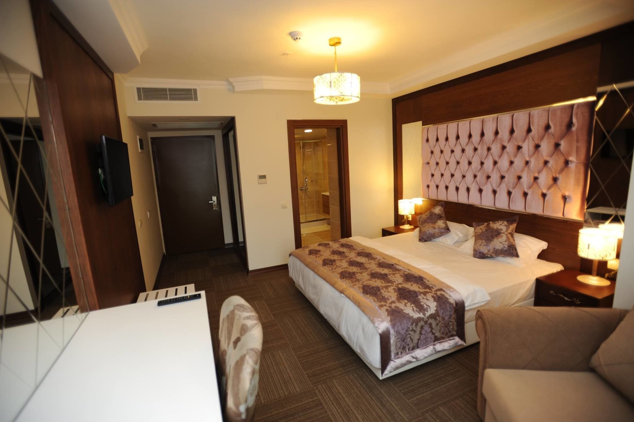 Una habitación de hotel con una cama grande, una mesita de noche con lámparas, una silla y un televisor colgado en la pared.