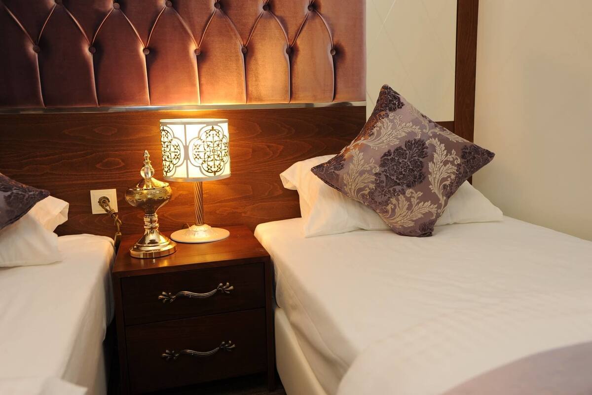 Una camera d'albergo con una testiera trapuntata, un comodino con una lampada, due cuscini e un mobiletto in legno con cassetti.