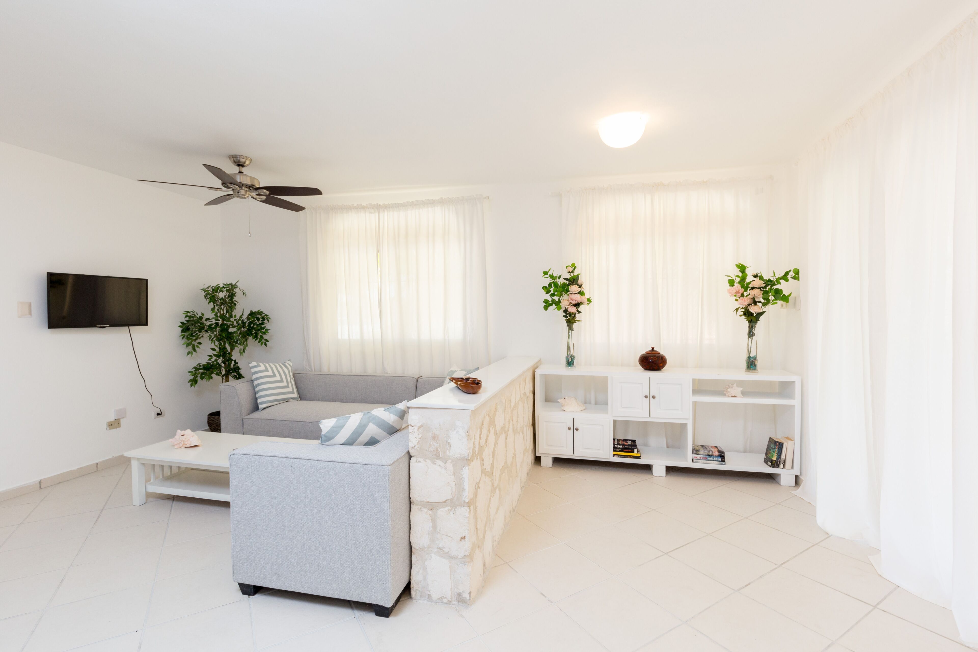Una sala de estar moderna con un sofá gris, una mesa de centro blanca y una pared de acento de piedra. Hay un ventilador de techo, un televisor de pantalla plana montado en la pared y un armario blanco con objetos decorativos.