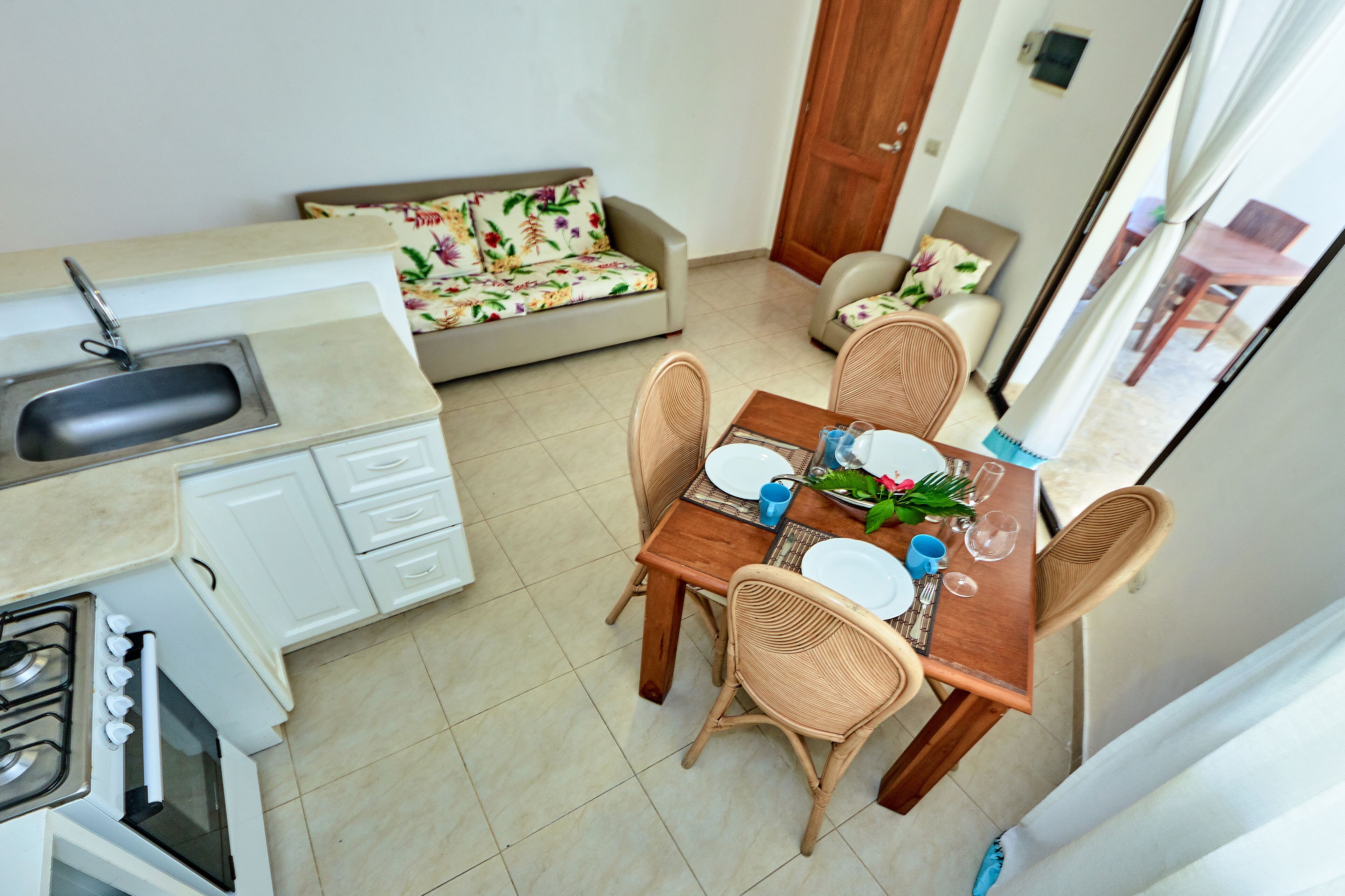 A compact kitchen with a sink, stove, and microwave, a dining area with a table set for two, and a sofa with a floral cushion.
