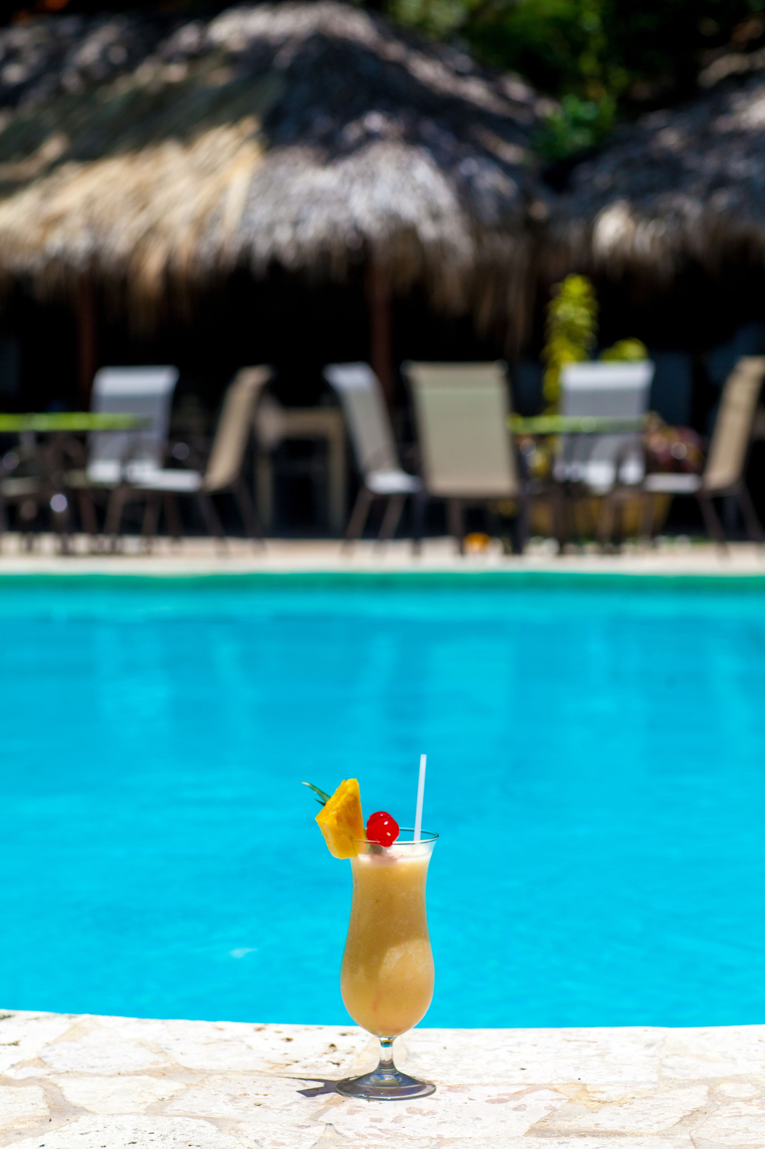 A cocktail with a straw and garnish in front of a pool with lounge chairs and a thatched umbrella in the background.