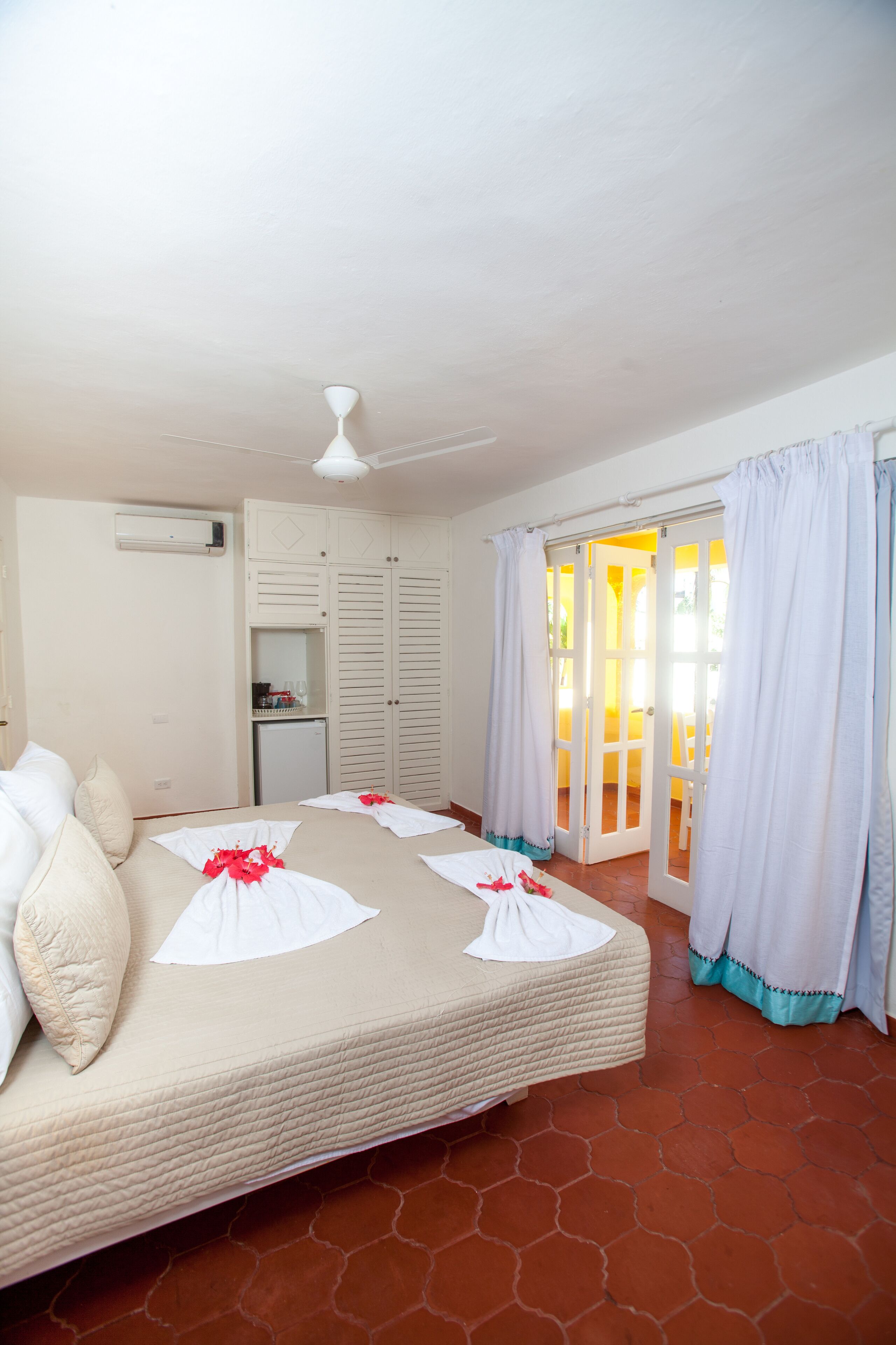 A hotel room with a bed, a ceiling fan, a minibar, and a sliding glass door leading to a balcony.