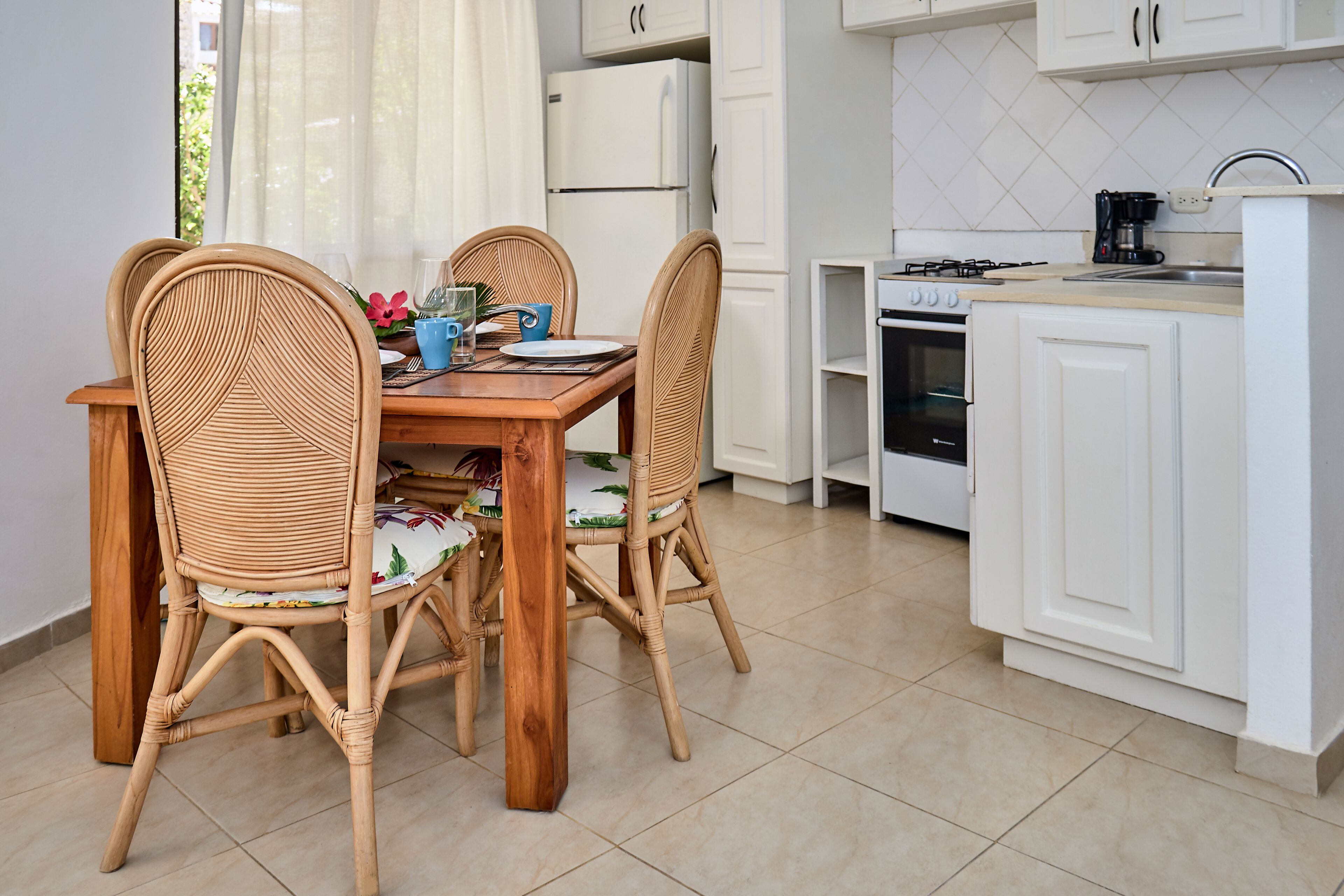 Una cocina compacta con armarios blancos, una mesa de comedor con sillas de mimbre y un horno pequeño.