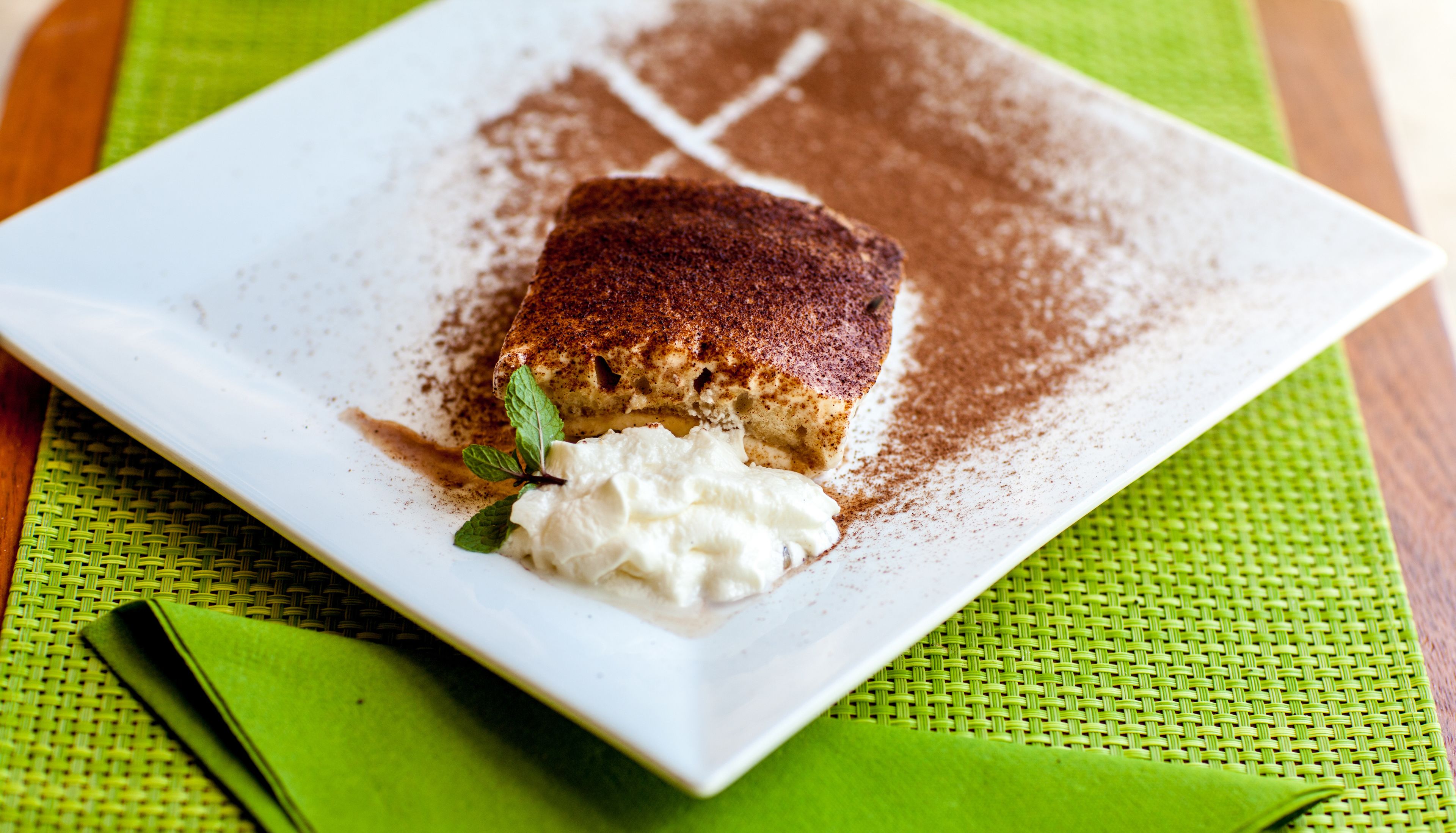 A square plate with a slice of cake topped with powdered sugar and a dollop of whipped cream, garnished with a mint leaf, on a green woven placemat.