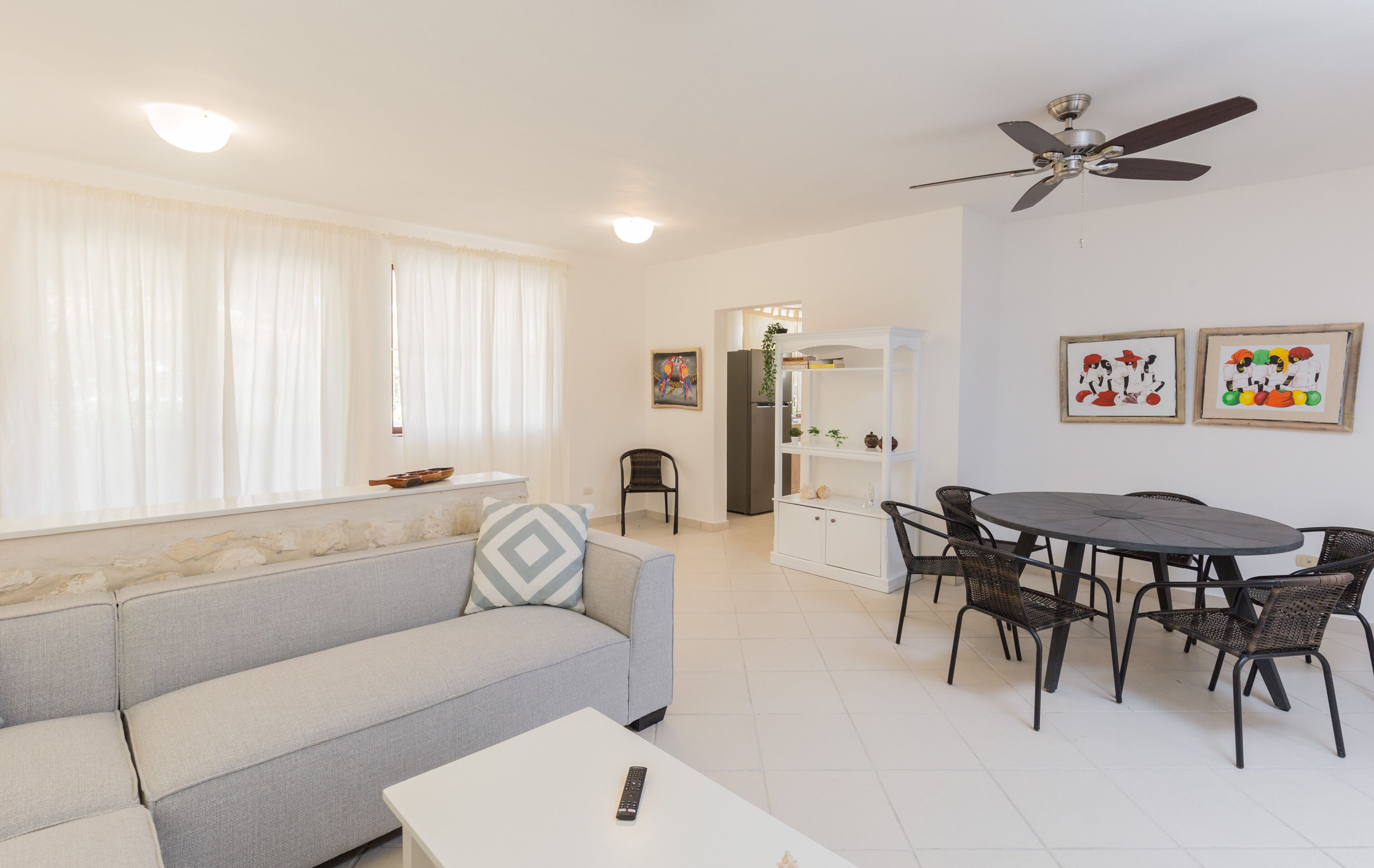 A living room with a sofa, a dining table with chairs, a ceiling fan, and a refrigerator.