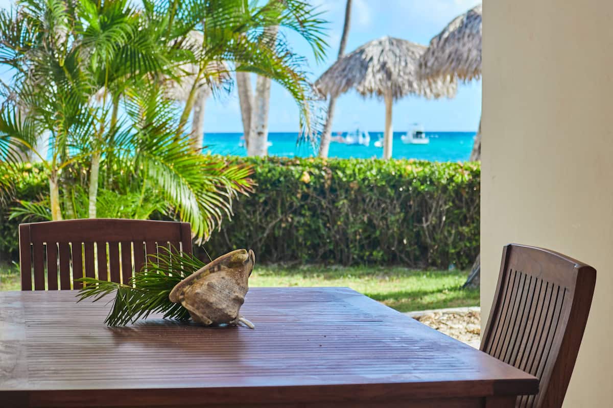 A wooden table with a shell and palm frond, set against a backdrop of a beach with palm trees and a thatched roof structure.
