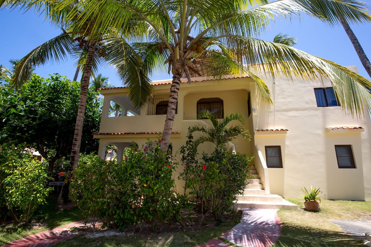 A two-story building with a balcony, surrounded by palm trees and various plants.