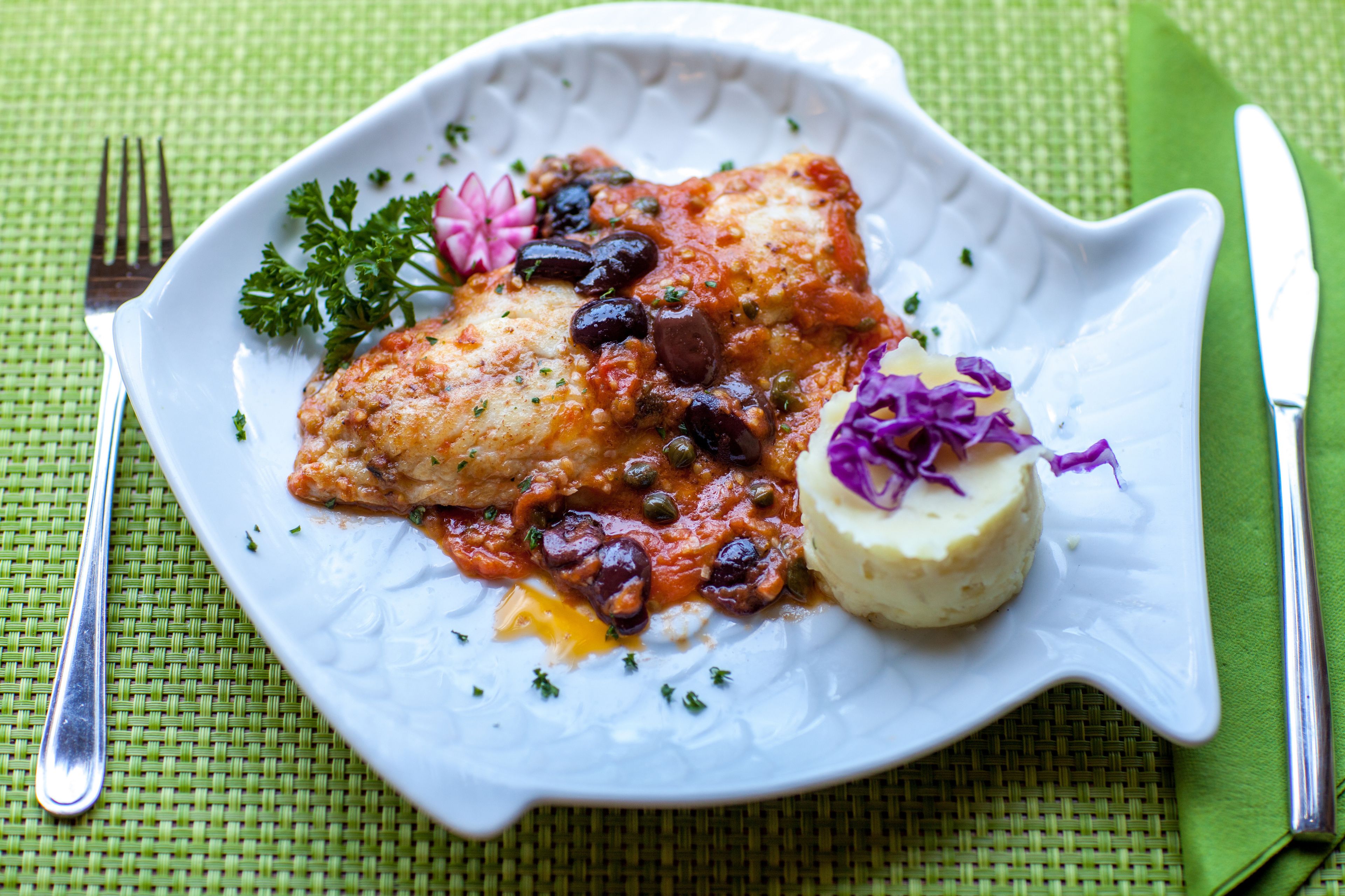 A white fish fillet with a tomato-based sauce, garnished with herbs and olives, served on a white plate with a decorative edge.