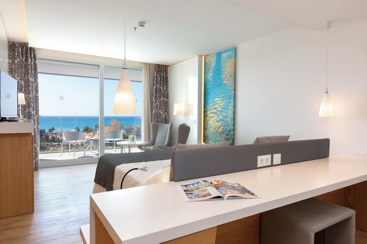 Une chambre d’hôtel moderne dotée d’une grande fenêtre, d’un coin repas, d’un canapé et d’un bureau avec un magazine.