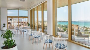 A spacious indoor area with large windows offering a view of the sea and buildings. There are several white chairs with blue cushions arranged in rows.