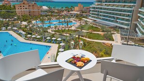 A balcony with a table set for a meal, overlooking a pool and resort area with buildings and a view of the sea.