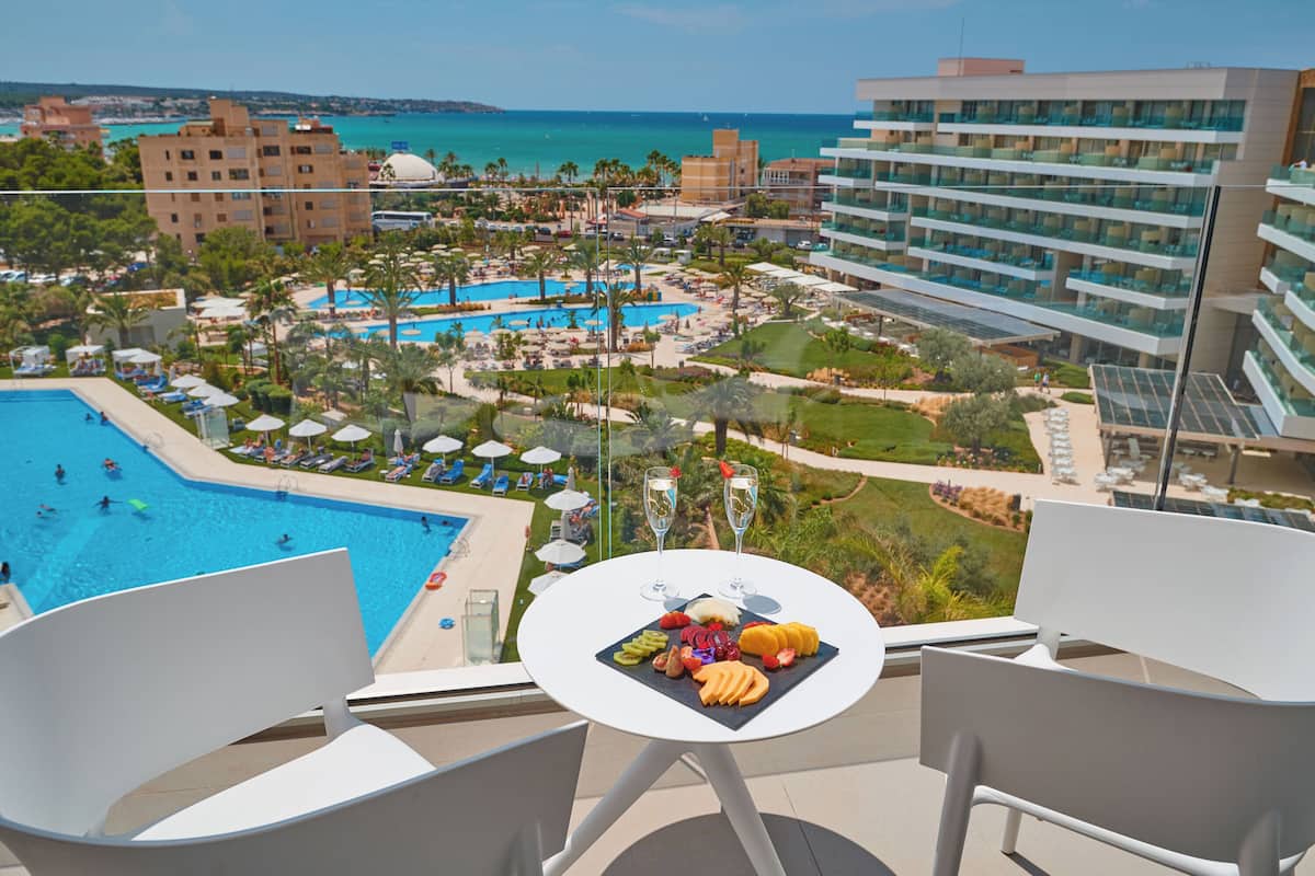 Un balcon avec une table dressée pour un repas, donnant sur une piscine et une zone de loisirs avec des bâtiments, et offrant une vue sur la mer.