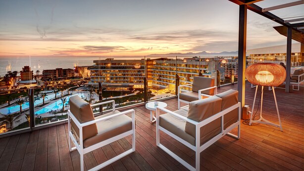 A balcony with white chairs and a table, overlooking a pool and a cityscape at sunset.