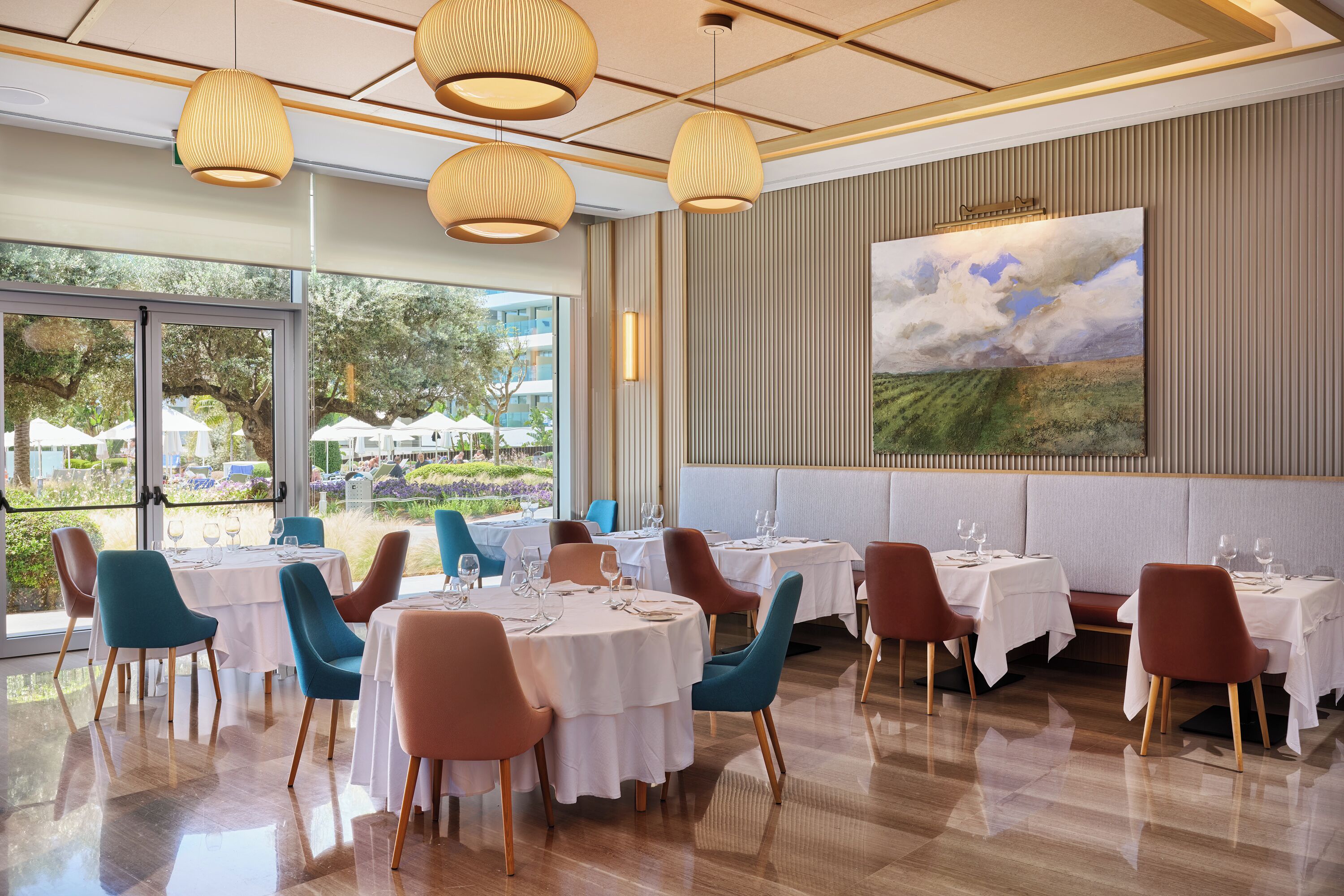 A restaurant with round tables, teal and brown chairs, and a large painting on the wall.