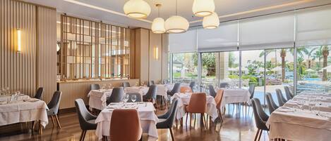 Ein Restaurant mit mehreren Tischen und Stühlen, großen Fenstern und hängenden Pendelleuchten.