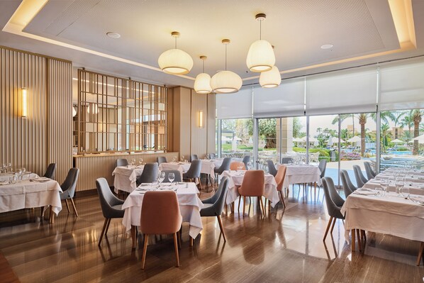 Un restaurante con varias mesas y sillas, ventanas amplias y lámparas colgantes.