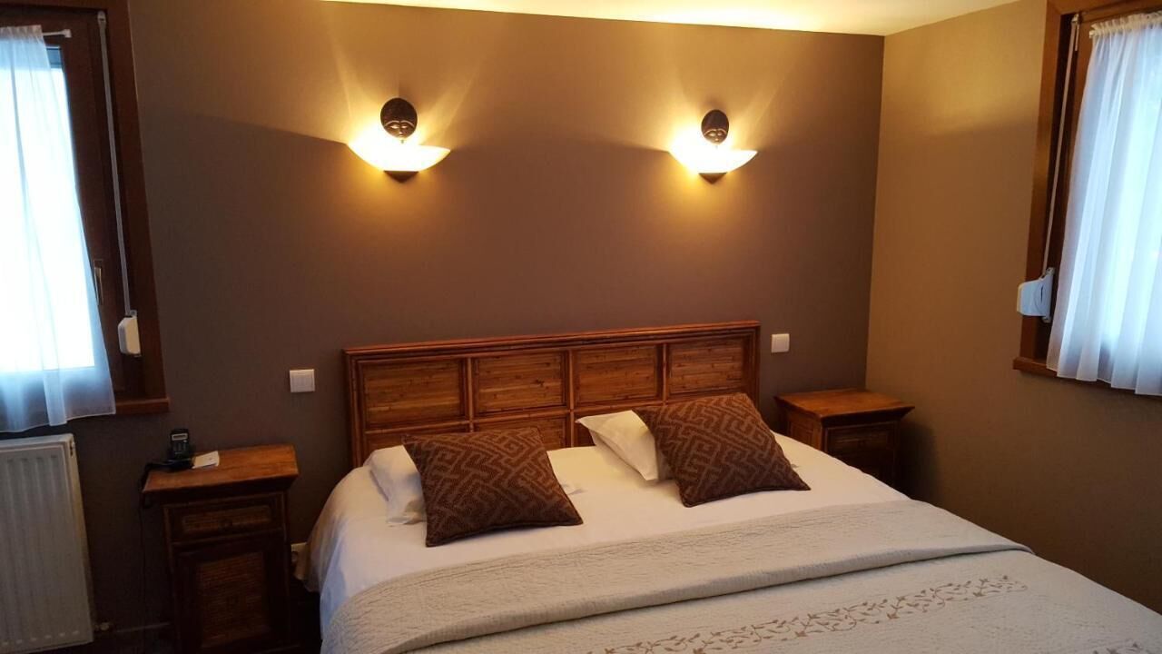Habitación de hotel con cabecera de madera, dos lámparas de noche, cama con ropa blanca y almohadas estampadas, y una ventana con cortinas.