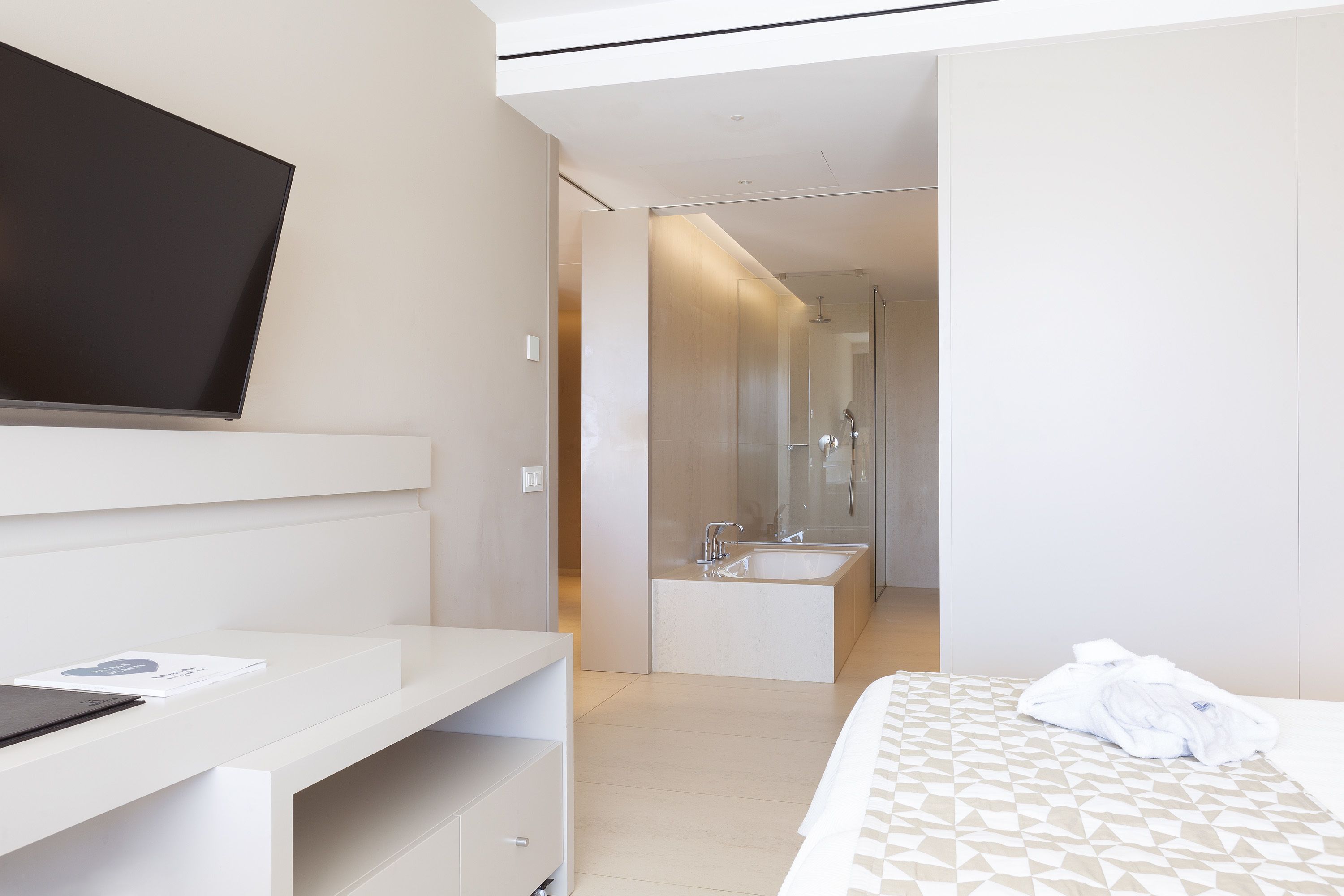 A modern hotel room with a flat-screen TV, a desk, a large bed, a bathroom with a shower, and a mirror.