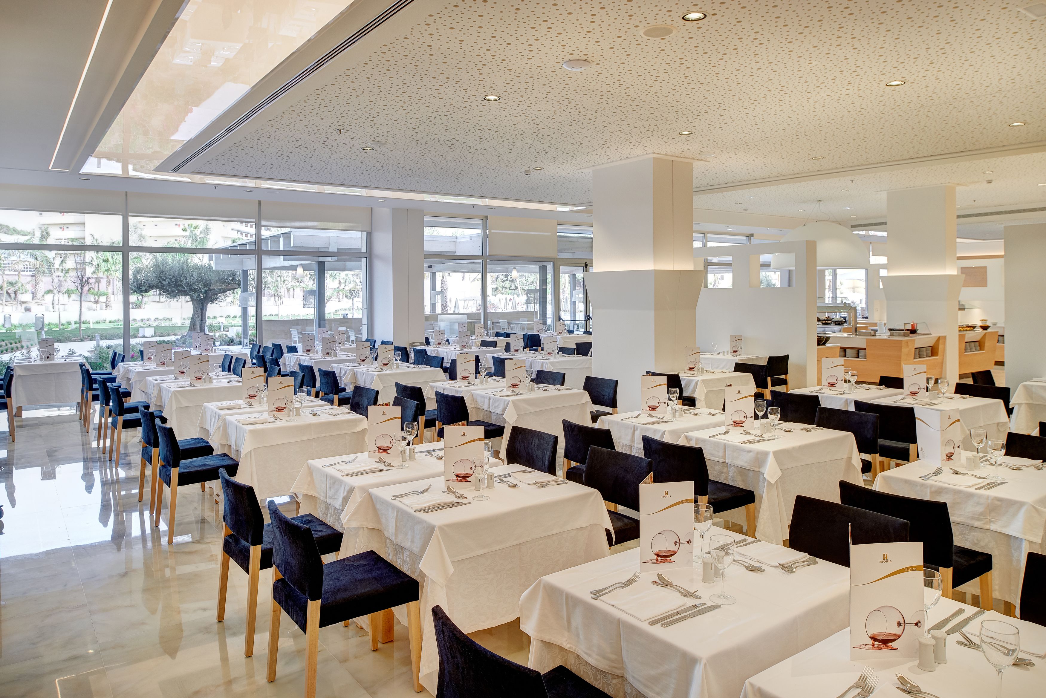 A spacious dining area with multiple tables and chairs, large windows, and a modern ceiling design.