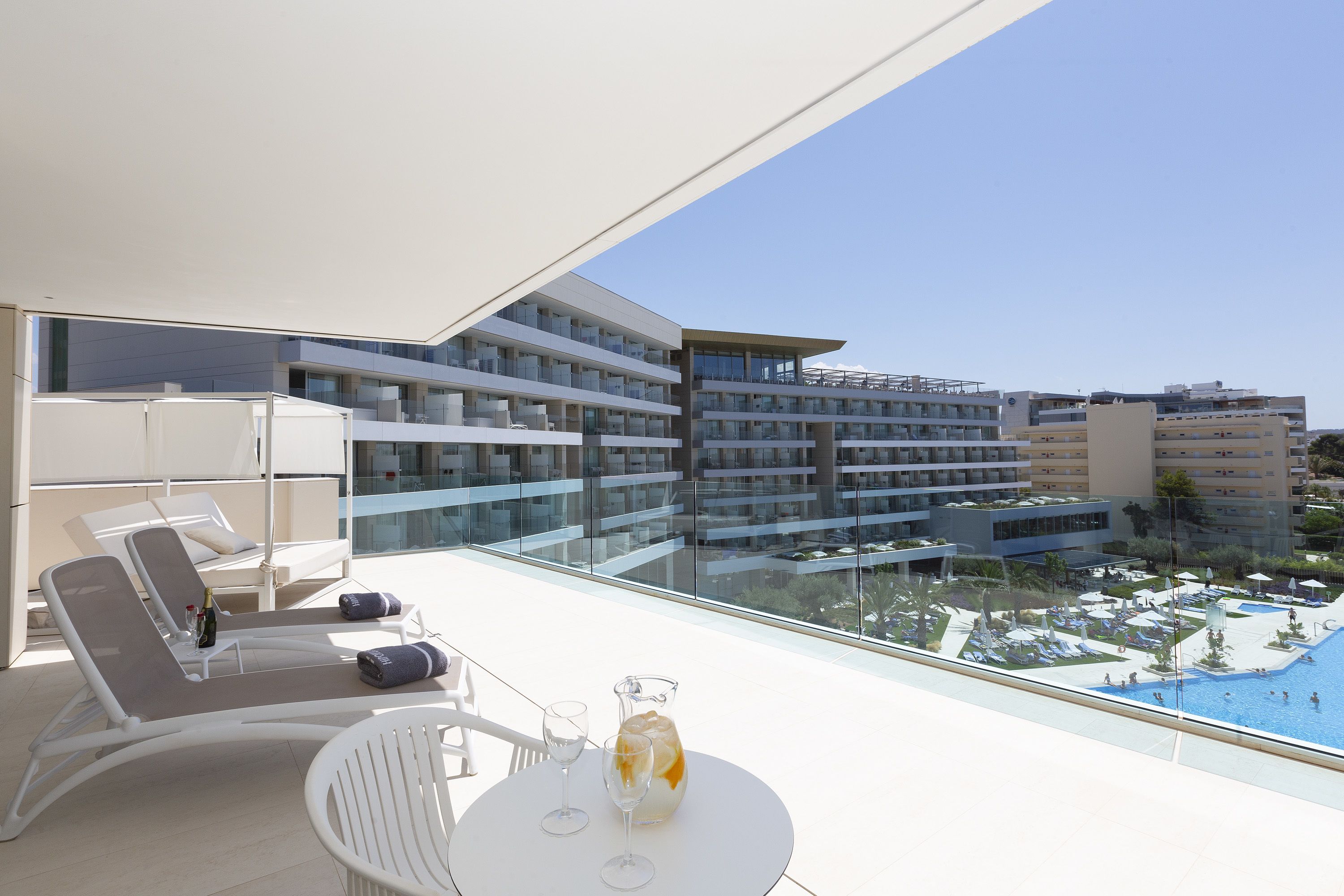 A modern hotel balcony with sun loungers, a table with drinks, and a view of the pool and surrounding buildings.