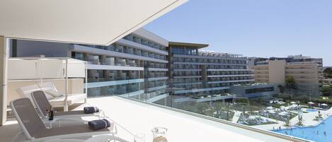 A modern hotel balcony with sun loungers, a table with drinks, and a view of the pool and surrounding buildings.