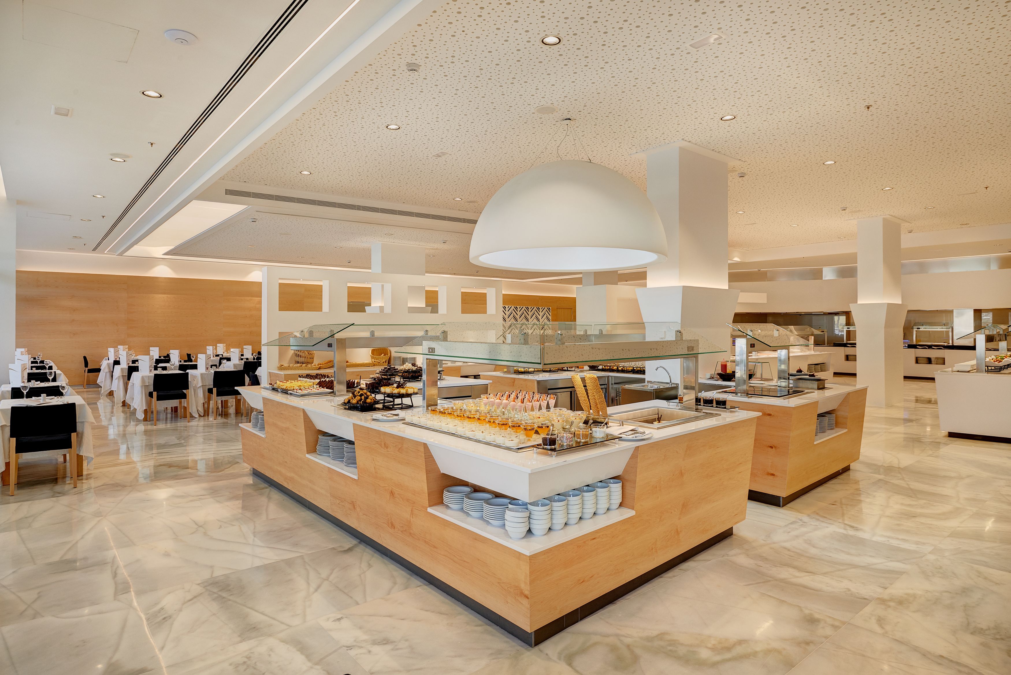 A modern hotel lobby with a central buffet area, multiple seating arrangements, and a spacious layout.