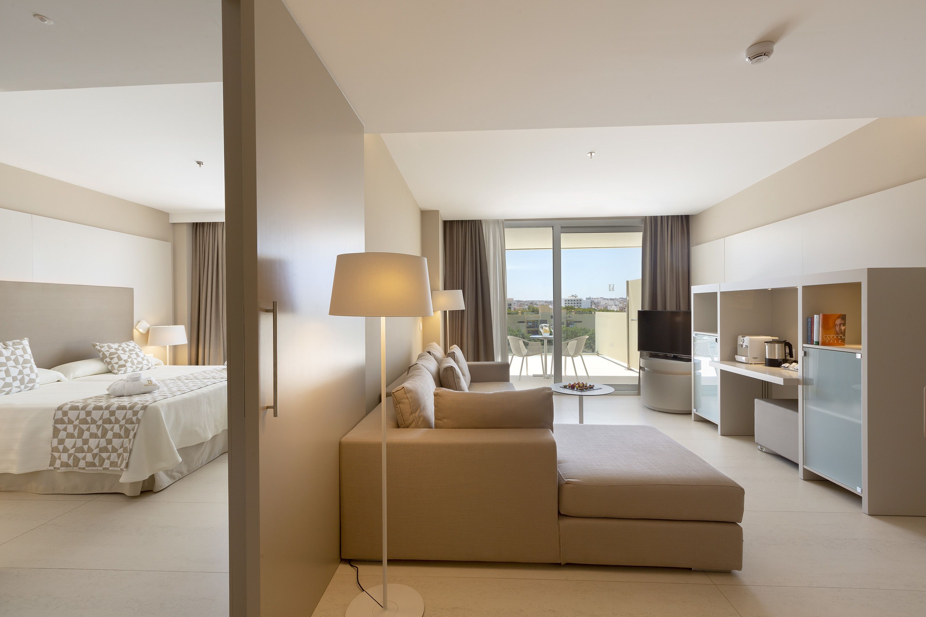 A modern hotel room with a bed, sofa, TV, and kitchenette.