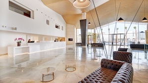 A spacious lobby with a high ceiling, a reception desk, seating area, and large windows offering views of palm trees and a parking area. - Hipotels Gran Playa de Palma (Palma de Mallorca)
