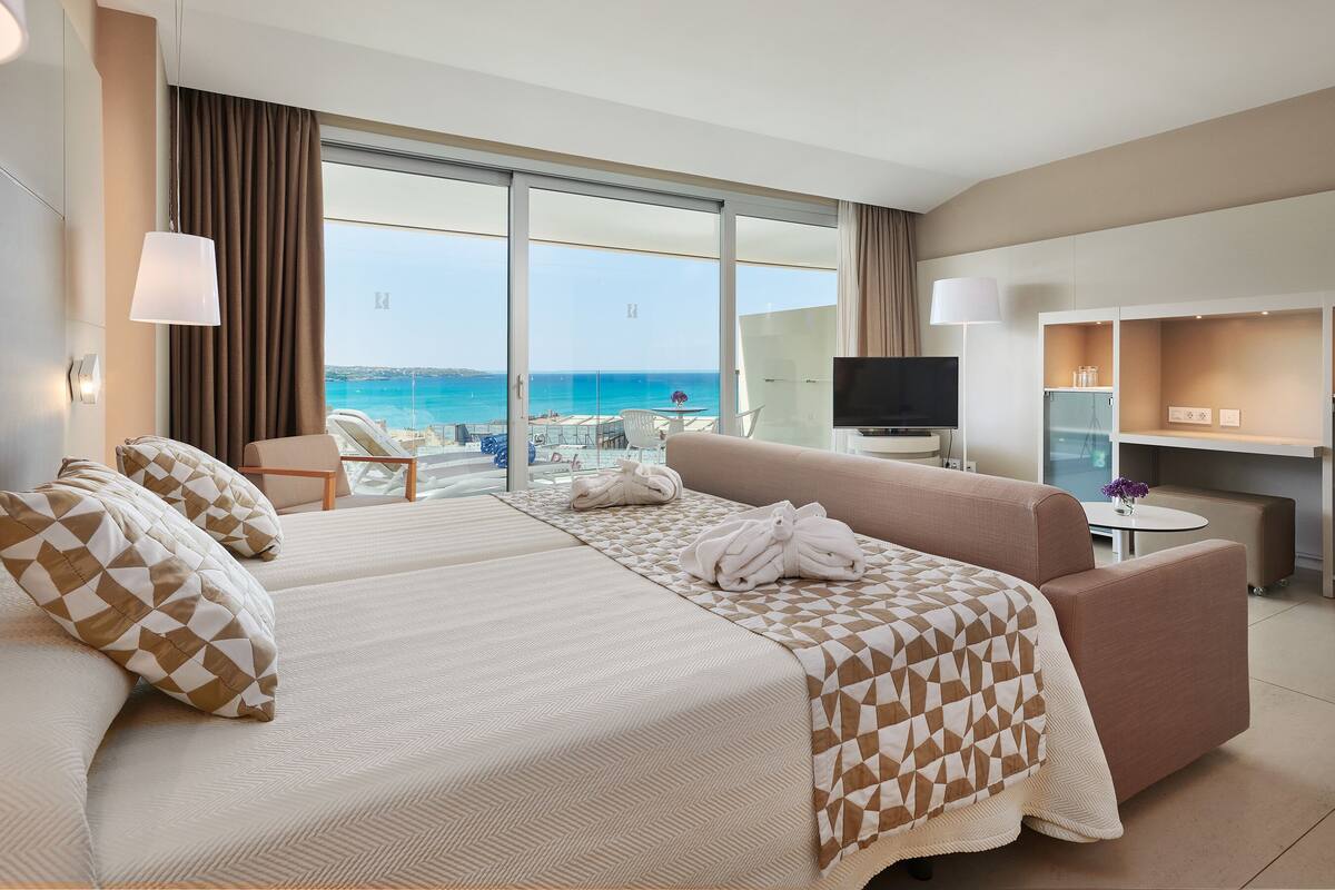 Quarto de hotel com cama, sofá, televisão e escrivaninha. Vista ampla da praia e do oceano através de grandes janelas.
