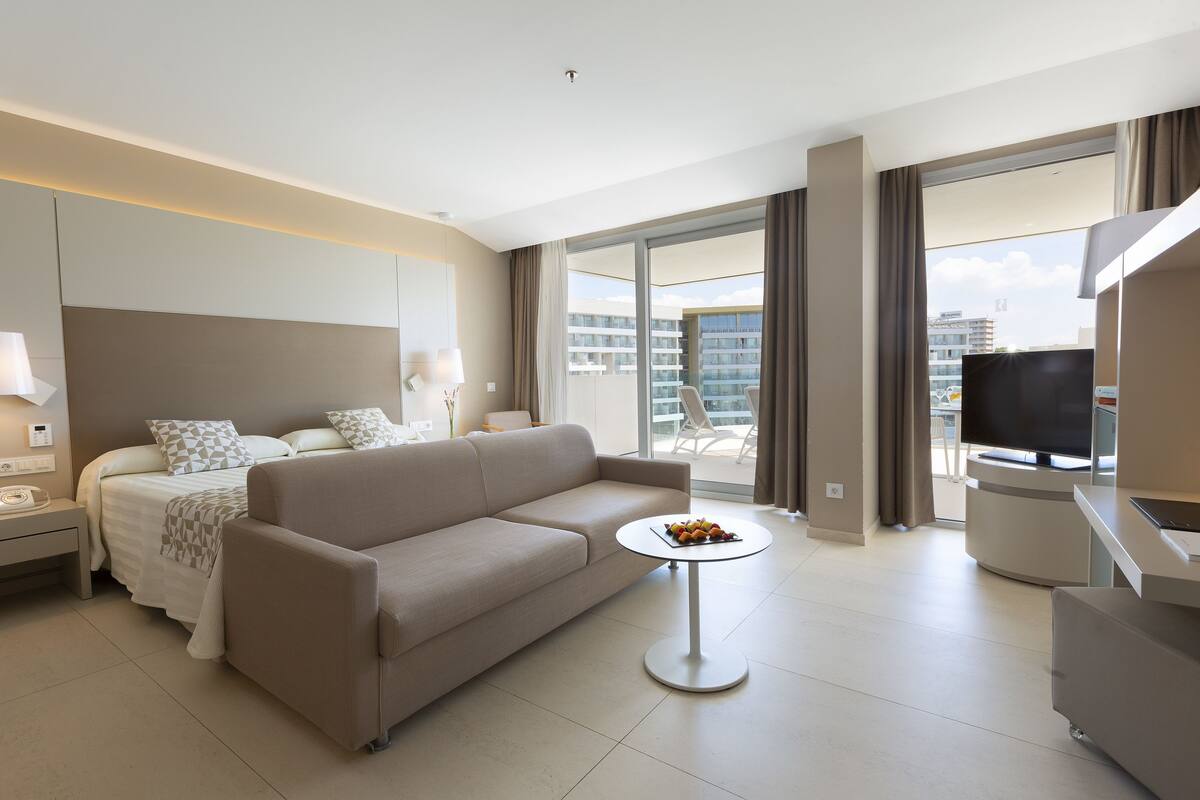 Quarto de hotel moderno com cama, sofá, televisão e varanda com vista para a cidade.