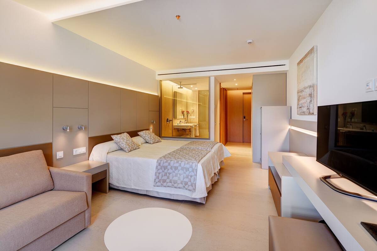 Quarto de hotel moderno com cama, sofá, televisão e banheiro visível através de uma porta de vidro.