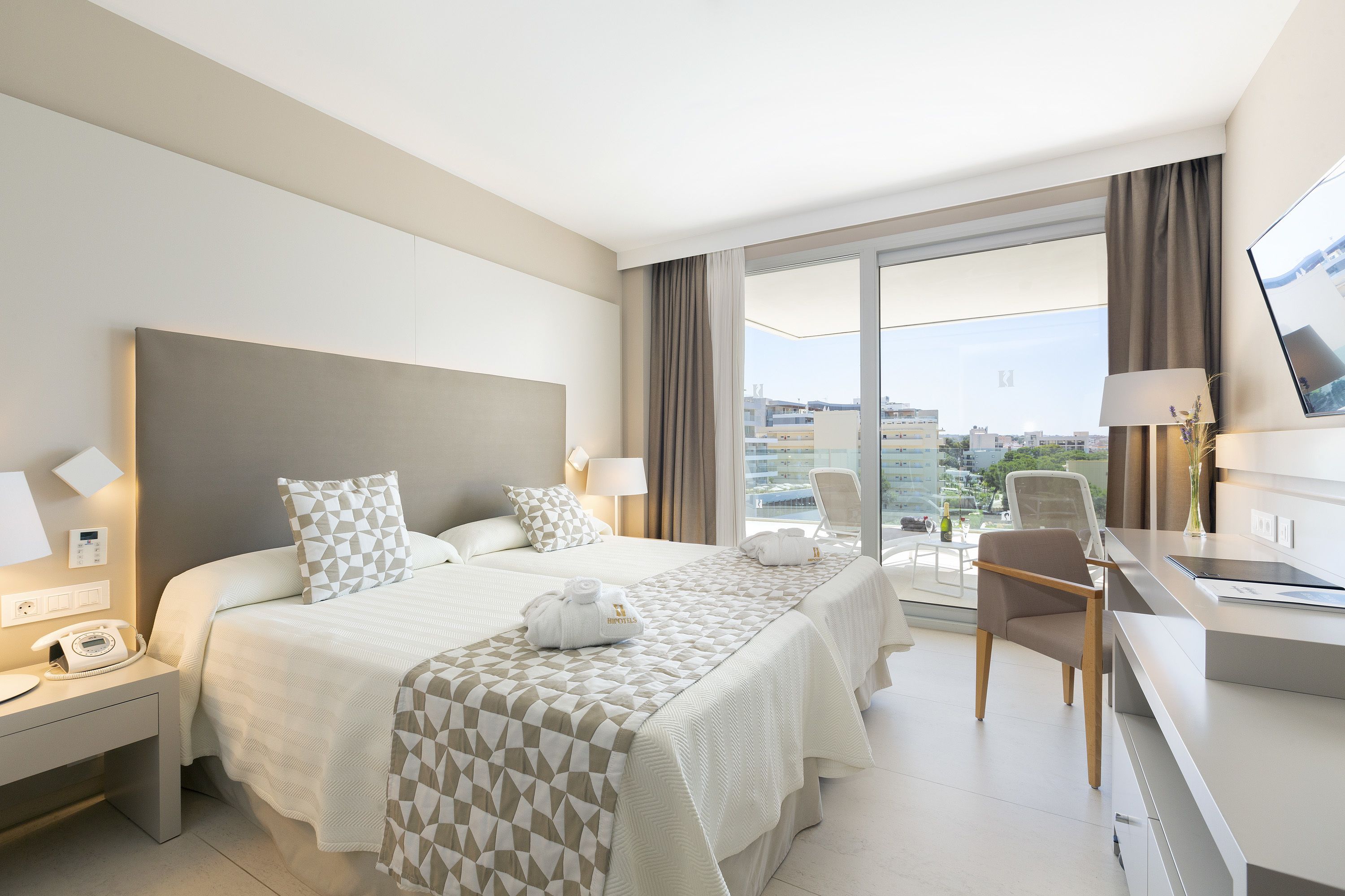 A hotel room with a bed, bedside tables, a desk, a chair, and a view of the city.