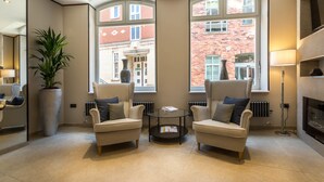 Un moderno salón con dos sillones, una mesa de centro, una planta grande en maceta y vistas a un edificio de ladrillo rojo a través de amplios ventanales.