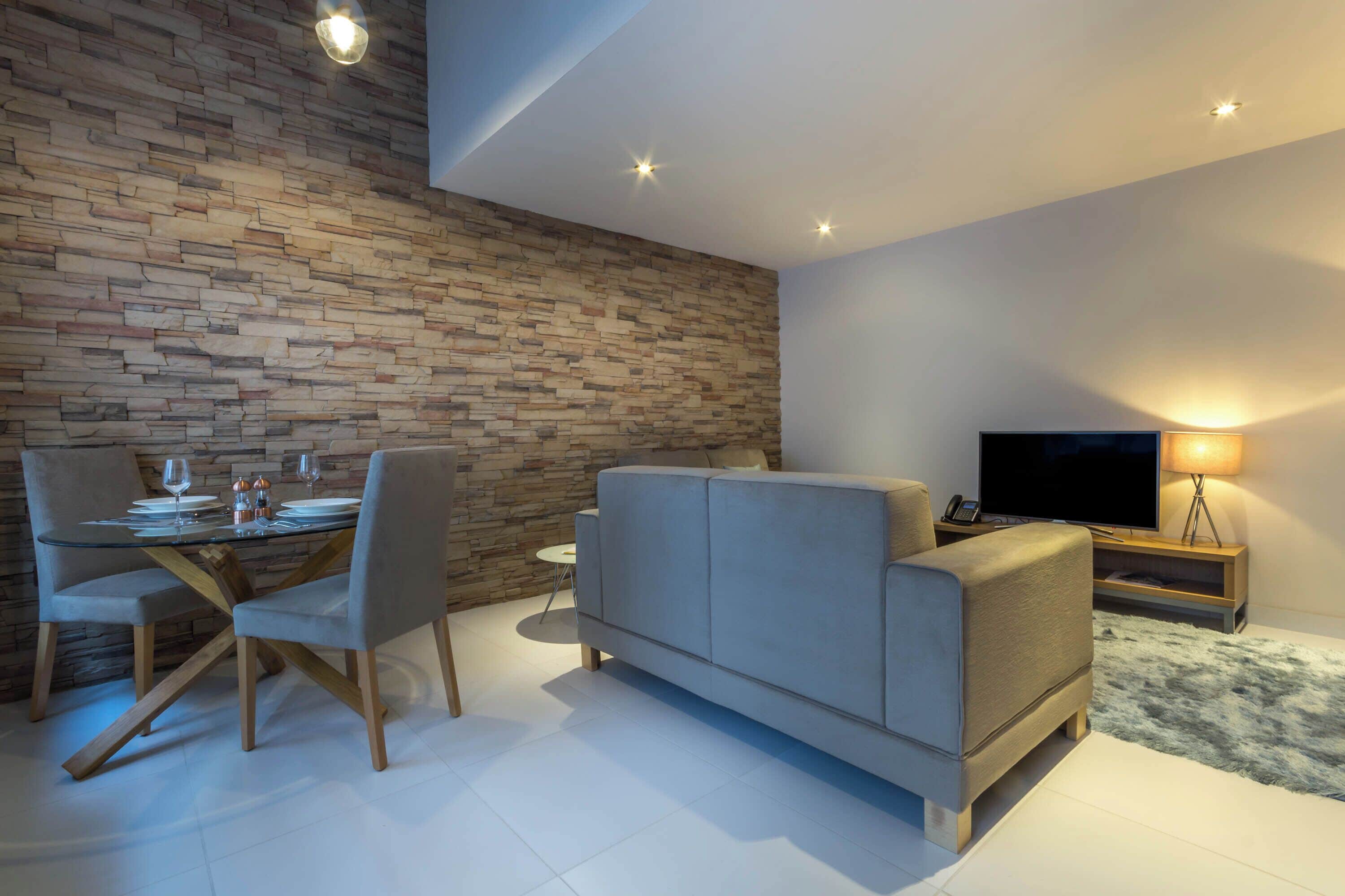 Ein modernes Wohnzimmer mit Steinwand, Glastisch, grauem Sofa und Fernseher auf einem Holzboden.