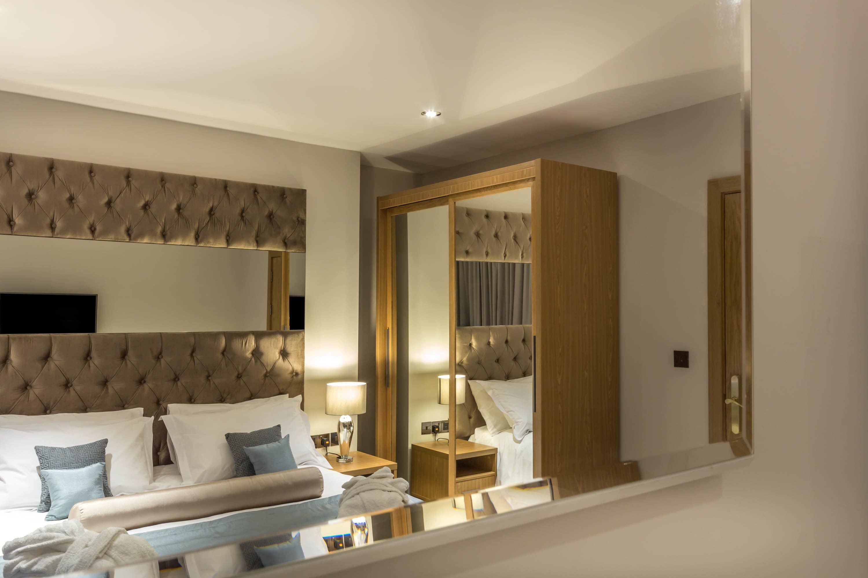 Een hotelkamer met een gestoffeerd hoofdeinde, een grote spiegel, een nachtkastje met een lamp en een keurig opgemaakt bed met kussens en een deken.