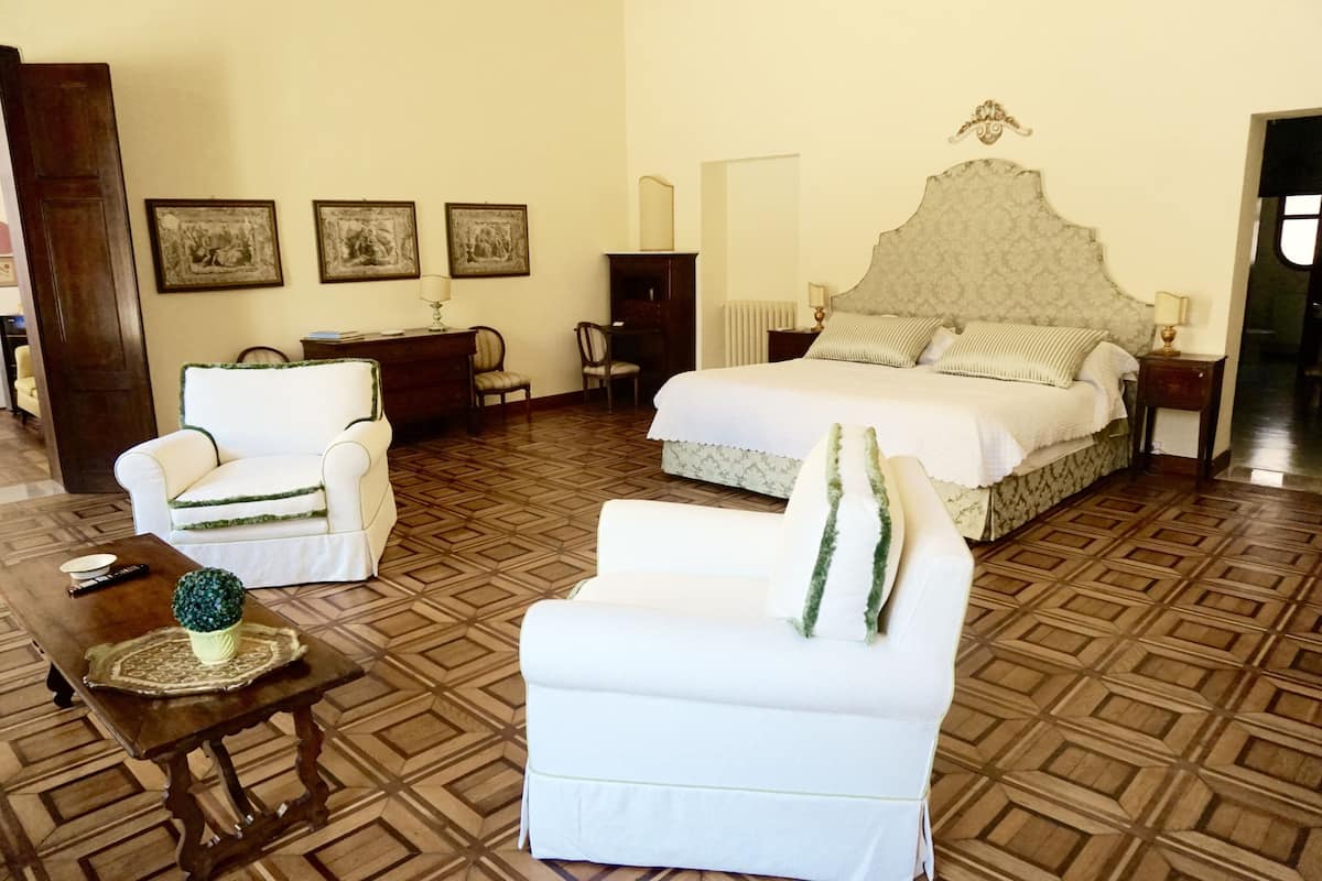 Una habitación de hotel con una cama grande, dos sillones blancos, una mesita de madera y una pequeña planta en maceta.