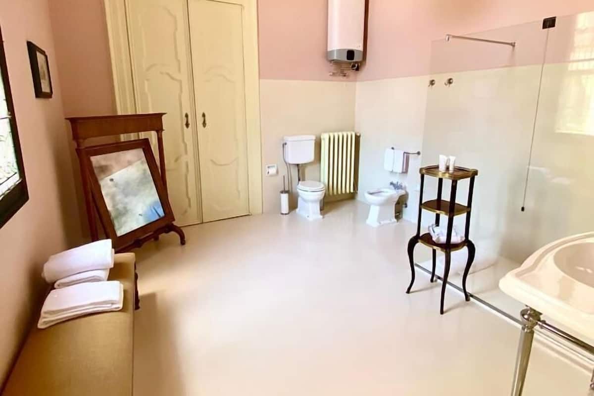 Baño con inodoro, bidé y bañera. Hay un perchero y un cuadro en un banco.