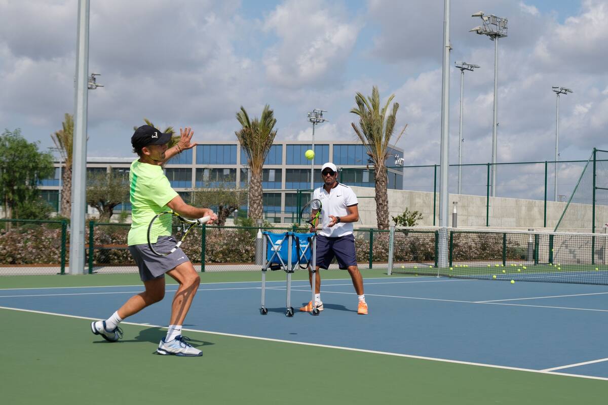 A tennis court with two players, a blue bag, and a building in the background.