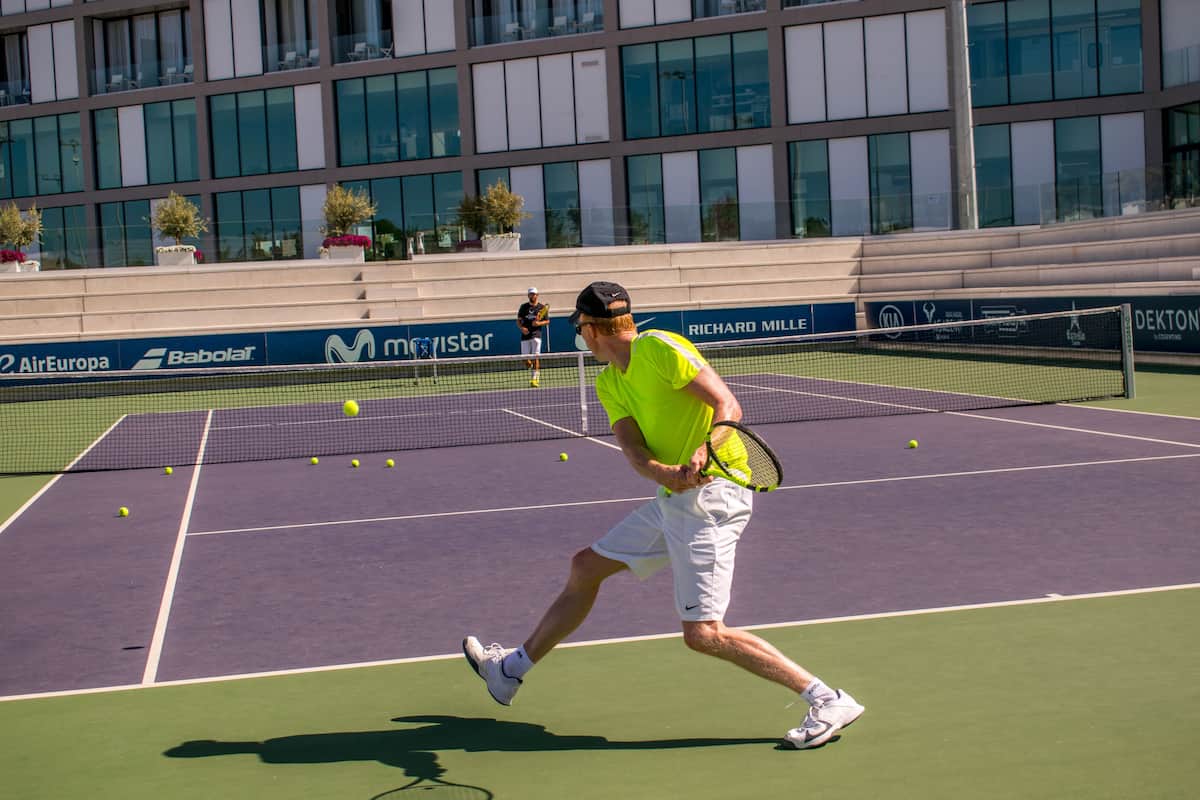 A tennis court with a net, a player in a bright yellow shirt, and a building with advertisements in the background.