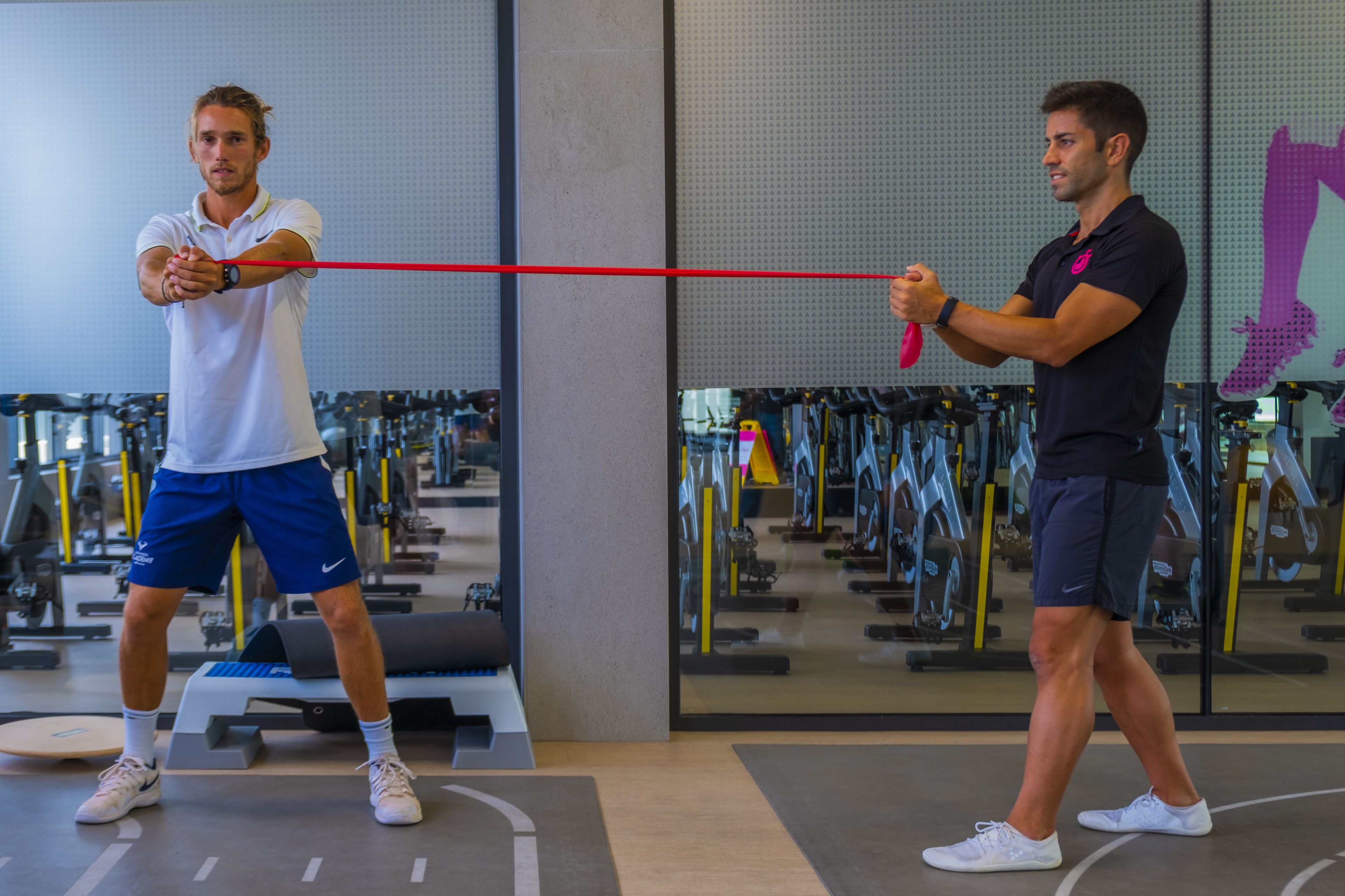 Two men exercising with resistance bands in a gym with various equipment.