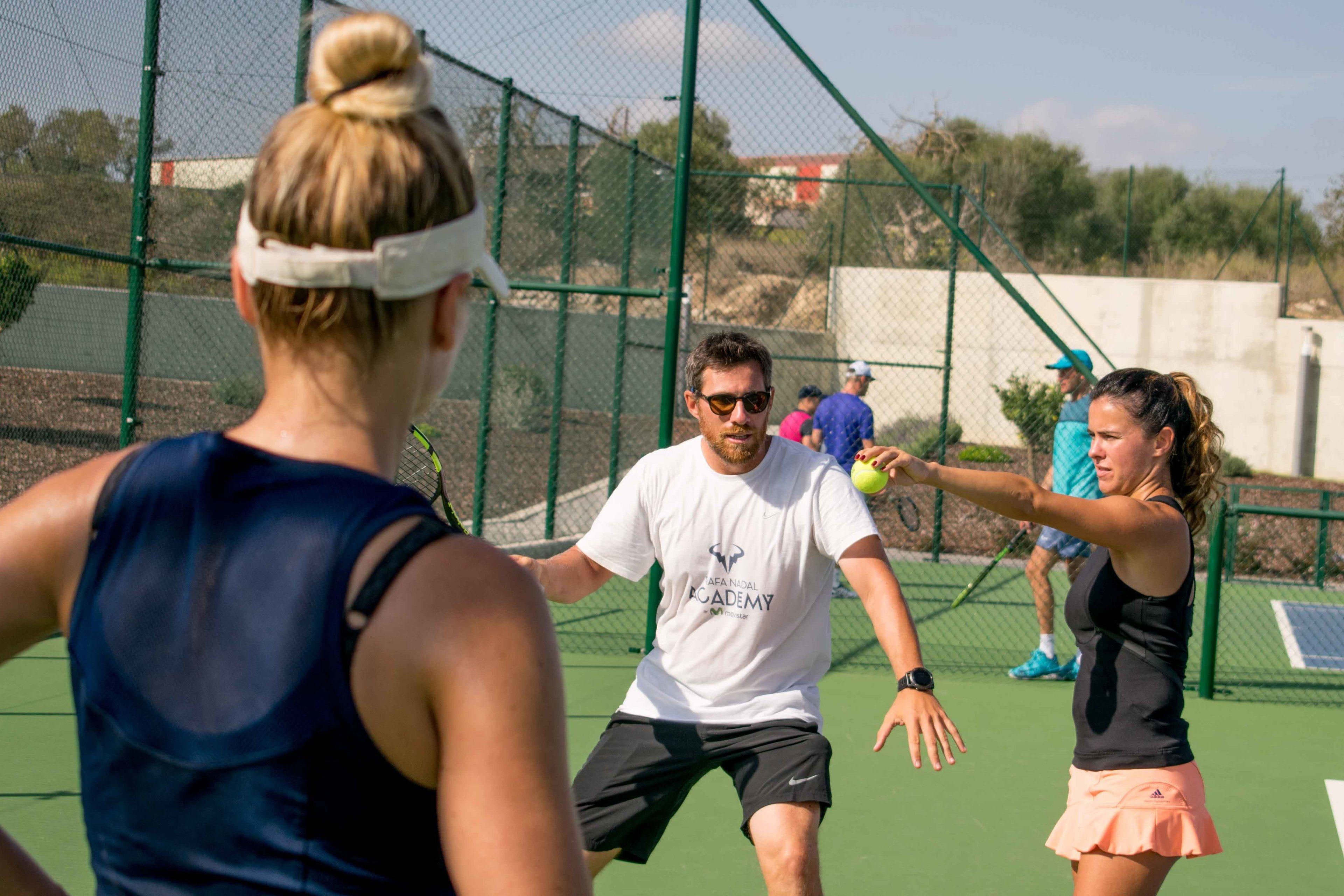 Una cancha de tenis con jugadores en movimiento, rodeada por una cerca de malla y follaje al fondo.