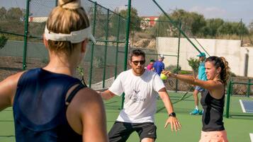 Una cancha de tenis con jugadores en movimiento, rodeada por una cerca de malla y follaje al fondo.