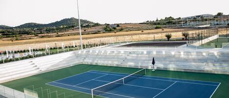 Una cancha de tenis exterior con gradas, un foco y vistas del paisaje circundante.