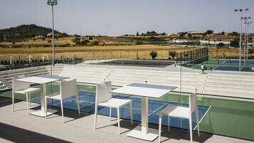Una cancha de tenis al aire libre con mesas y sillas blancas, rodeada por una valla y vistas a un paisaje montañoso.