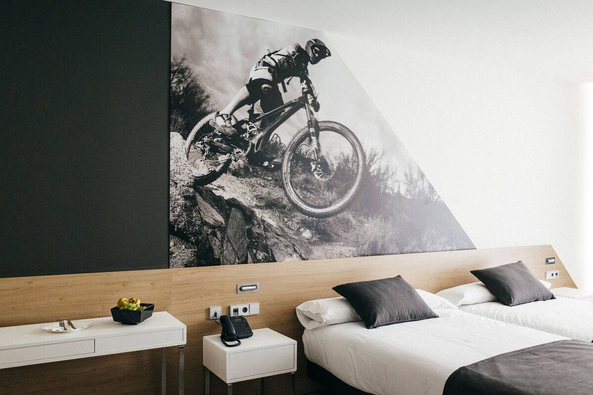 A hotel room with a bed, bedside tables, a small desk, and a large wall mural of a mountain biker.