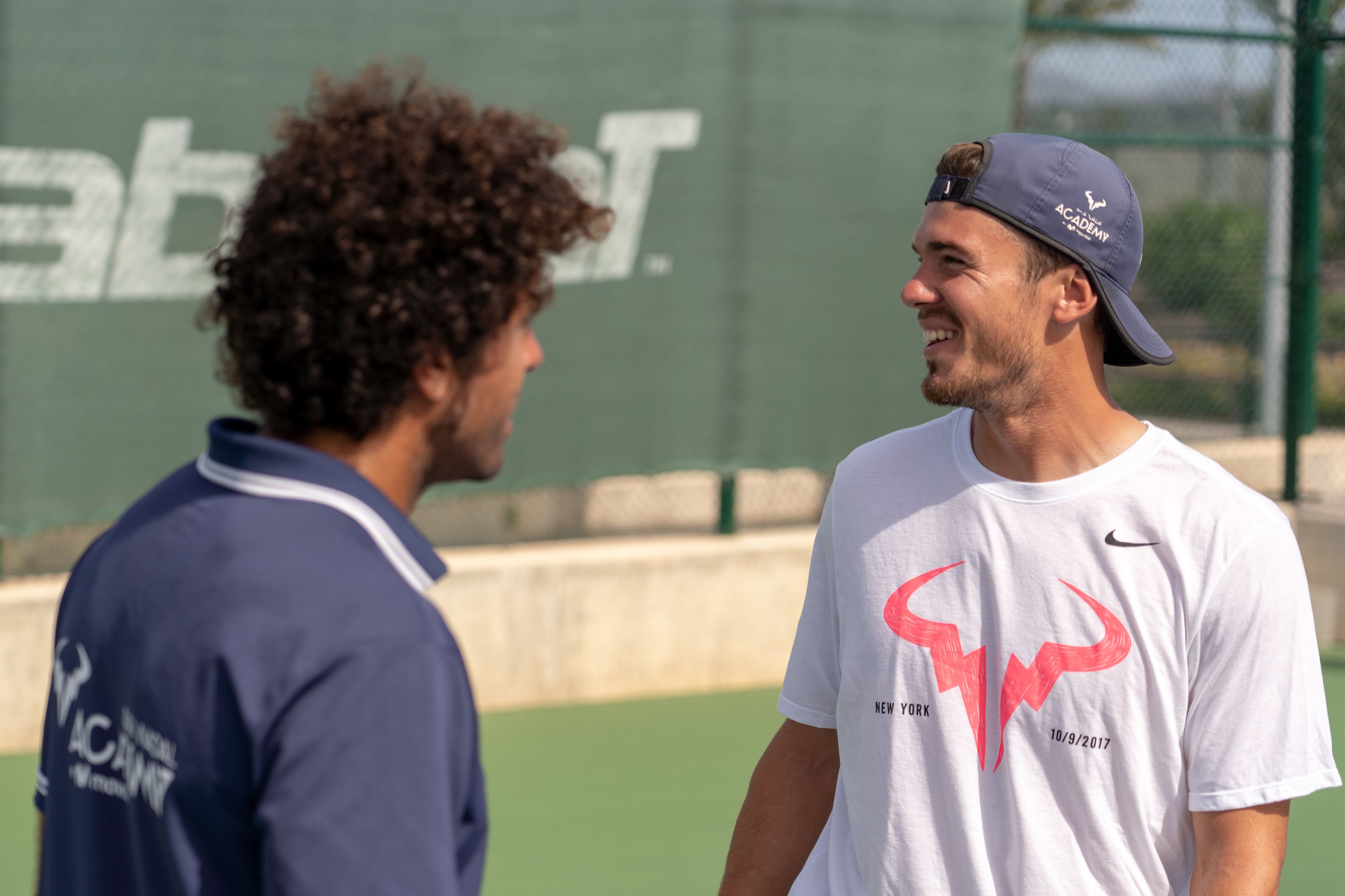 Two people on a tennis court with a green wall in the background.